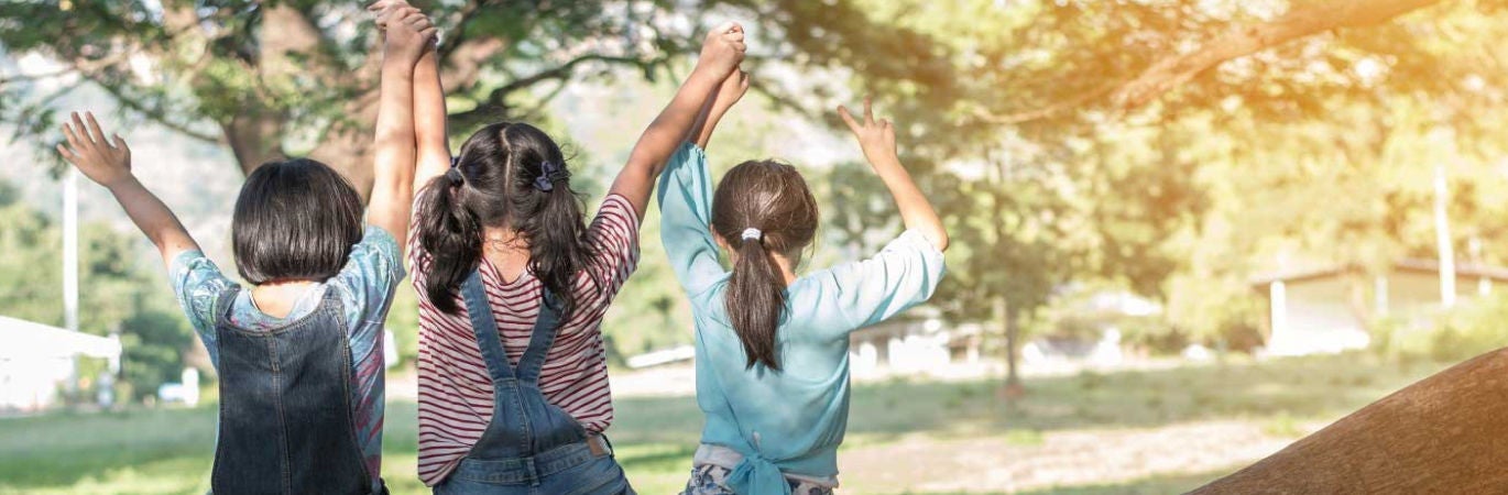 Three young girls sitting facing away from the camera on a tree branch, holding their hands in the air